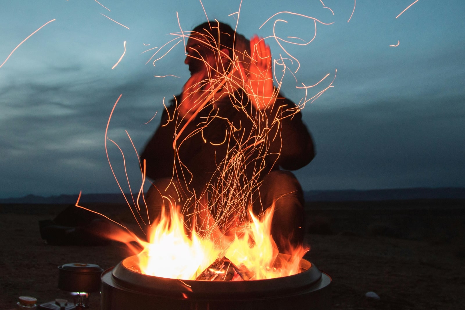 A person seated by a campfire, surrounded by flying sparks, creating a warm and inviting atmosphere in the night.