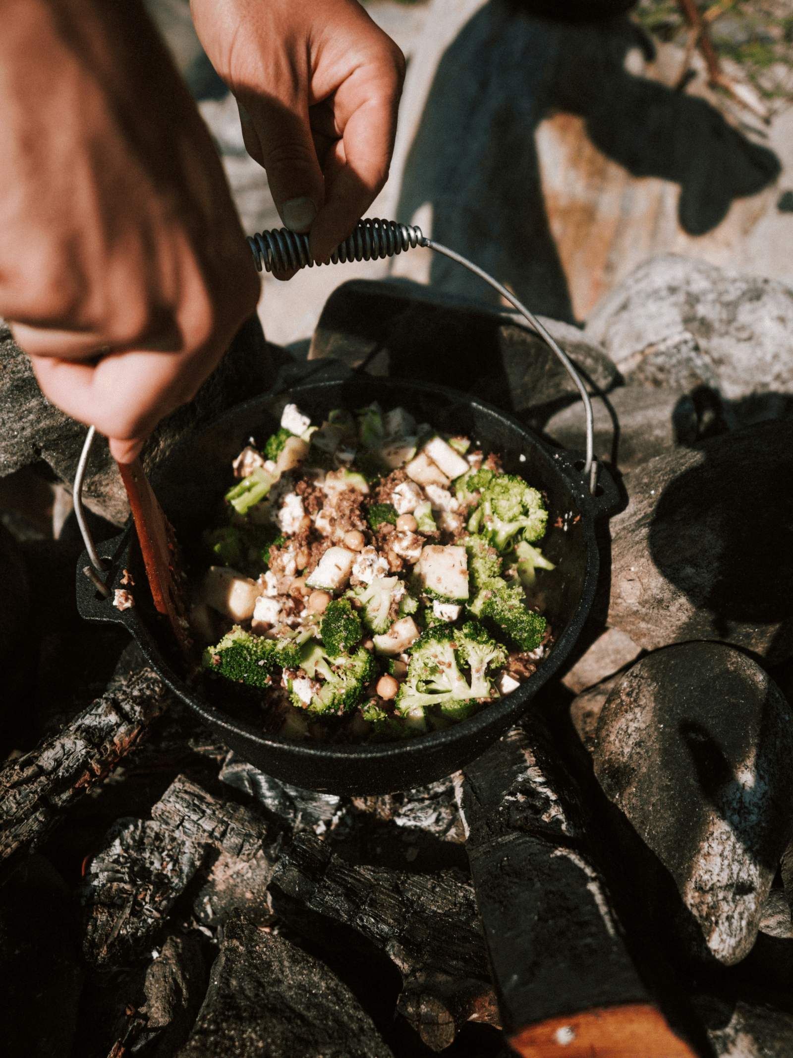 A person skillfully cooks food in a pan over an open fire, showcasing culinary techniques in a vibrant outdoor setting.