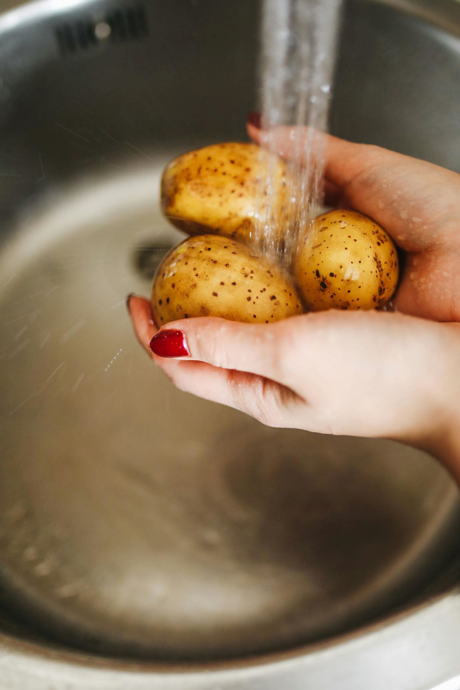 A person scrubbing potatoes in a kitchen sink, water flowing and soap bubbles visible around the vegetables.