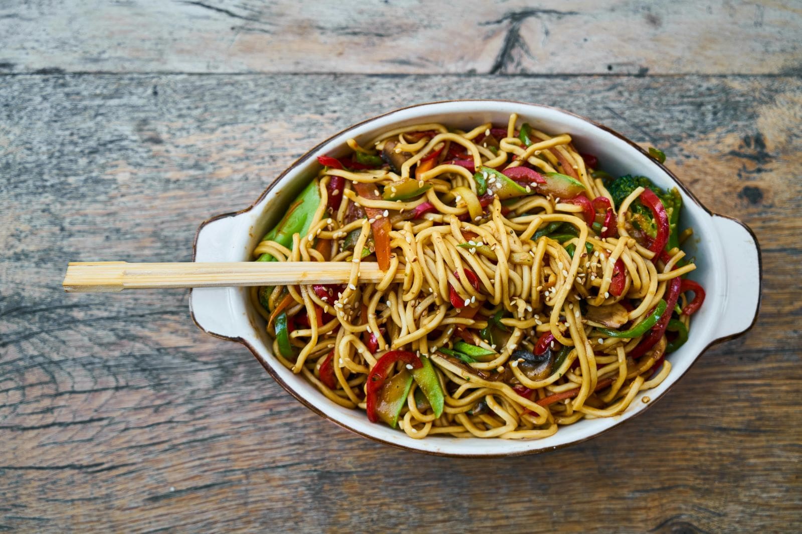 A bowl of noodles topped with fresh vegetables, accompanied by a pair of chopsticks resting on the side.