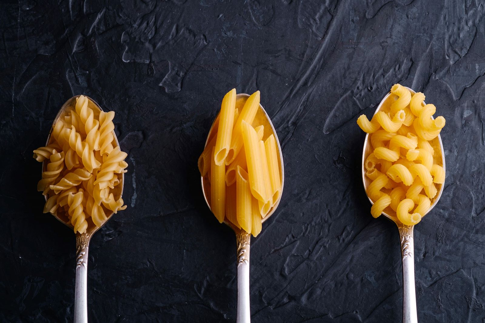 Three spoons each holding a different type of pasta, showcasing a variety of shapes and textures in a culinary display.