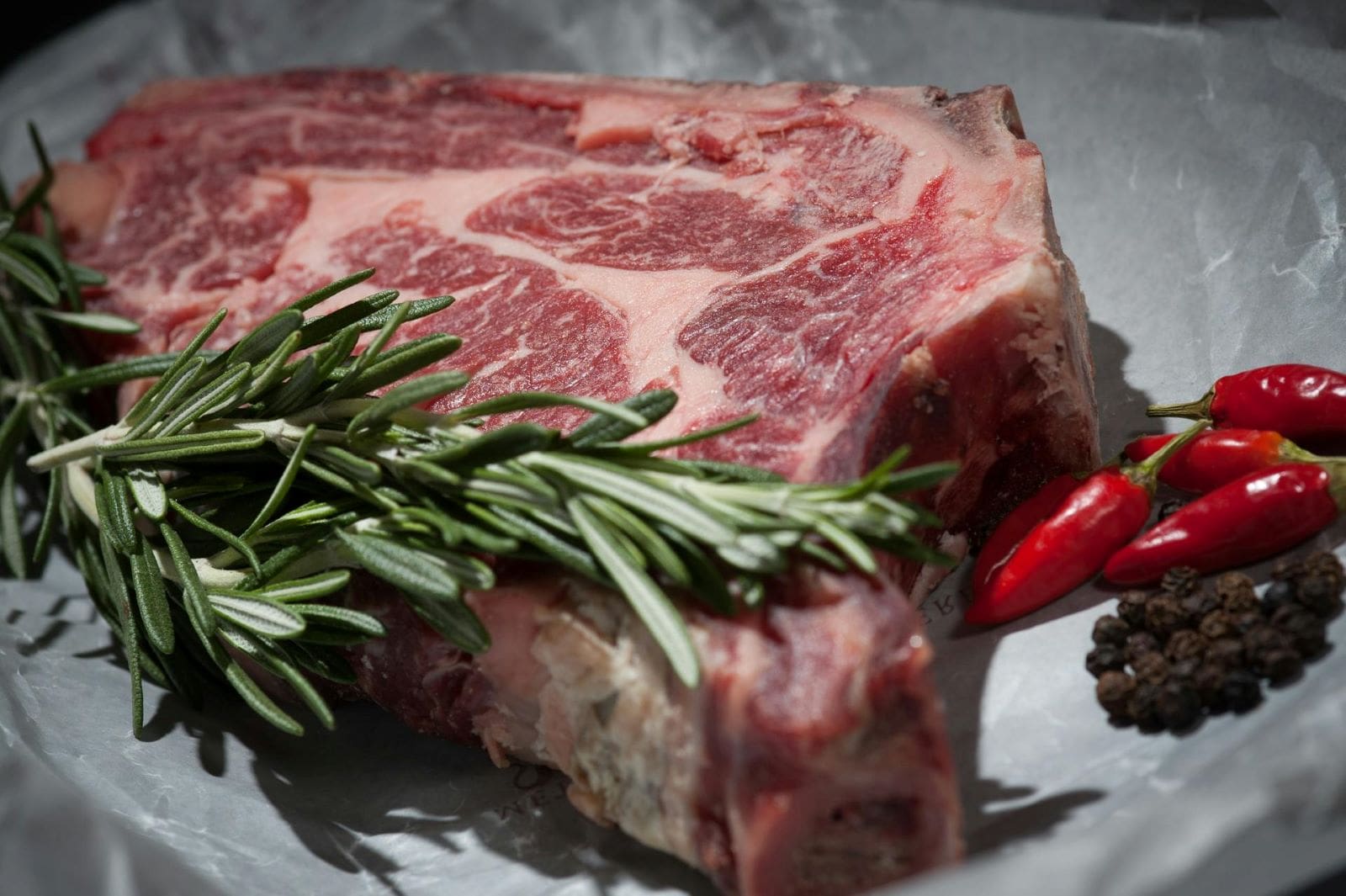 A raw beef steak garnished with fresh rosemary and sprinkled with black pepper, ready for seasoning and cooking.